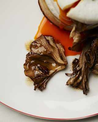 Close-up of sautéed mushrooms next a crumpet topped with a poached egg that spills vibrant orange yolk onto the white plate.