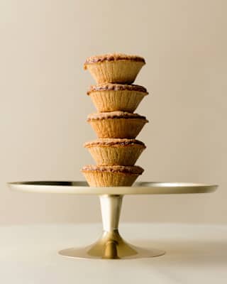 Close-up photograph of five baked pastry mince pies stacked in a tower on a silver cake stand, seen from the side.