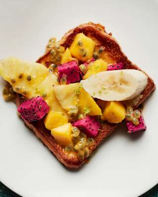 A slice of toast topped with pieces of banana, dragon fruit, pineapple, mango, and passion fruit sits on a white plate.