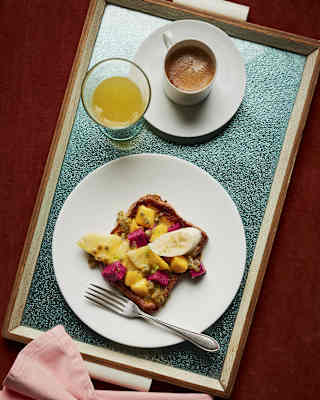 A plate with toast topped with banana slices and fruit, a fork, a cup of coffee, and a glass of orange juice on a tray, beside a folded pink serviette.