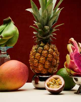 A pineapple stands upright among a mango, passion fruit, dragon fruit, lime, and a green leaf, all arranged against a red background.