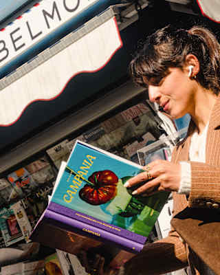 Low-angle image of a dark-haired woman reading Campania, with a painting of vegetables on a blue cover, by a newspaper stand.