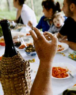 A bottle of wine in a raffia cooler rests on the edge of a dining table, where a family is enjoying a Mediterranean lunch.