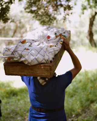 Walking through trees, a woman in a blue shirt carries a wooden box on her head containing her harvest, covered with cloth.