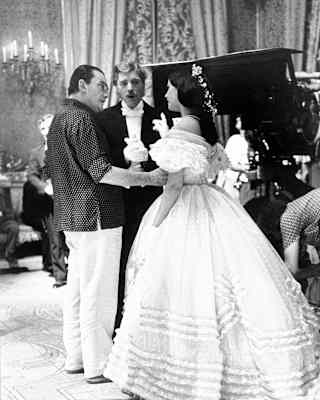 Visconti talks to Claudia Cardinale, who wears a white frilled dress, watched by Burt Lancaster on a film set, in monochrome.