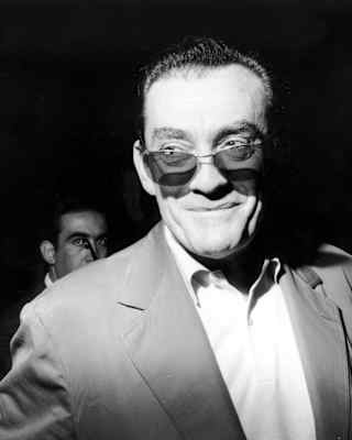 Film director, Luchino Visconti smiles as he is snapped in a suit with dark glasses, in a black and white close-up.