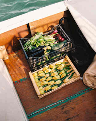 A waterborne boat carries two crates in its hull, one with zucchinis and flowers, the other with red onions, seen from above.