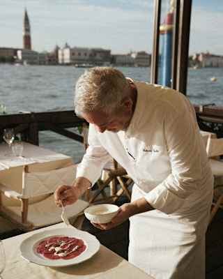 Chef Roberto Gatto, wearing chef's whites, dresses a plate of thinly-sliced bresaola at a Cip's Club waterside dining table.