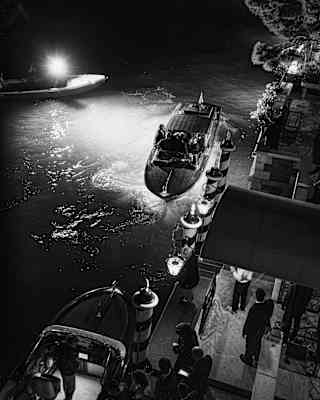 In darkness, hotel staff wait as a late boat pulls up alongside the striped paline posts, guided by lamps, seen from above.