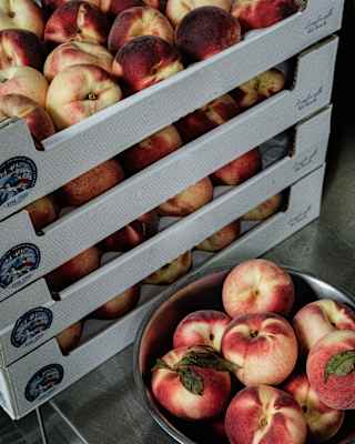 Beautiful Italian white peaches with red blushes fill a stainless steel bowl on a counter, alongside a stack of boxed peaches.