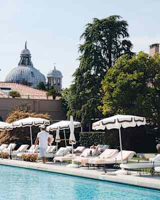 White sun loungers and parasols with brown-trim line the azure swimming pool, with the imposing dome of Le Zitelle behind.