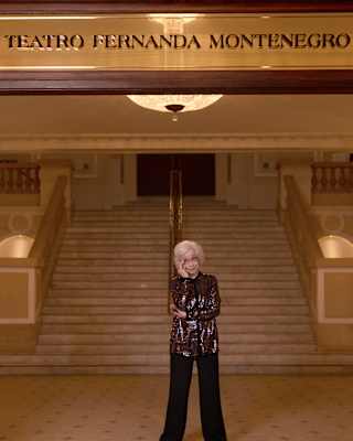 Fernanda Montenegro, with short grey hair and glasses, stands in front of a theatre in her name, wearing a dark sequined top.