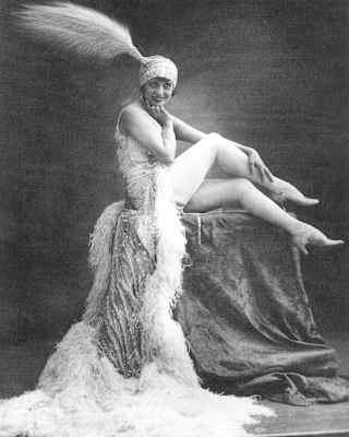 Black and white photograph of Moulin Rouge Burlesque star, Mistinguett, posing on a pedestal in a feather-edged gown and hat.