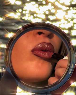 The lower face of a woman applying red lipstick is caught in a round compact mirror, with sea behind, taken by Sam Youkilis.