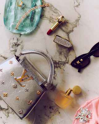A silver Louis Vuitton handbag with a gold logo, accompanied by a lipstick, perfume bottle, sunglasses, and jewellery on a marble surface.