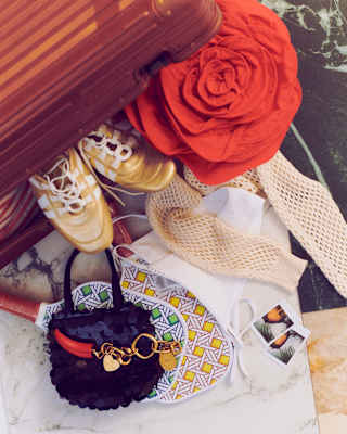 A suitcase filled with a red fabric flower, gold trainers, a white mesh top, patterned tennis racquets, a small sequinned bag with a chilli charm, and two polaroid photos.