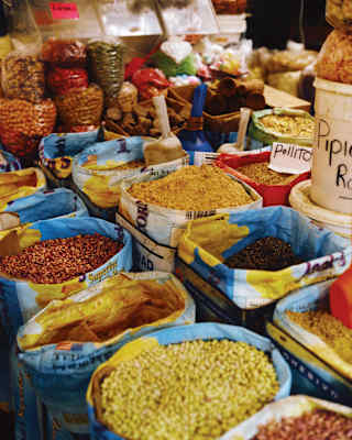 Bright image of two rows of open sacks containing grains and spices, and bags of dried beans, seen at a Mexican market stall.