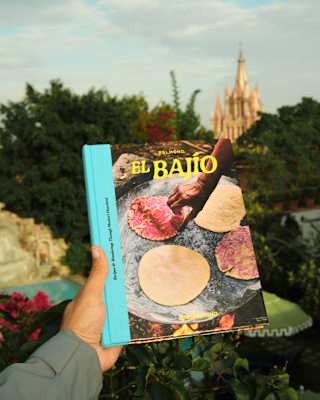 A hand holds the El Bajío book, with plain and red tortillas on the cover, in front of a view of San Miguel's iconic church.
