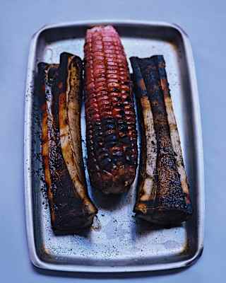 An cob of grilled red corn sits between two charred bones, with marrow exposed, on a silver cooking tray, seen from above.