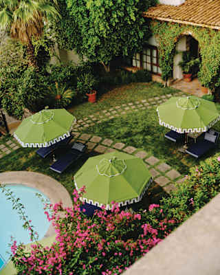 A courtyard garden with green parasols, lounge chairs, a small pool, stone pathways, and lush plants surrounding a building with a tiled roof and arched windows.