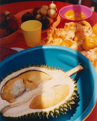 Close-up of a large durian fruit half in a blue bowl on a red table with bright plates of local Malay side dishes.