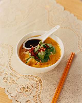 Looking down on a bowl of Assam Laksa fish and noodle soup, with a green garnish, chilli flakes and a spoon shrimp paste.
