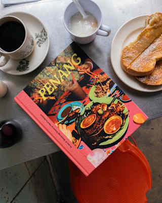 A copy of the Penang cookbook, with coral binding, rests on a breakfast table with black coffee and toast, seen from above.