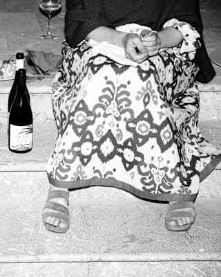 Seen waist down in black and white, two women in party dresses relax on marble steps with bottles of wine and buffet food.