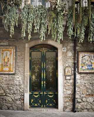 Trailing plants spill from a balcony above the ornate entrance to a ceramics chop with a painted double door and tile-art.