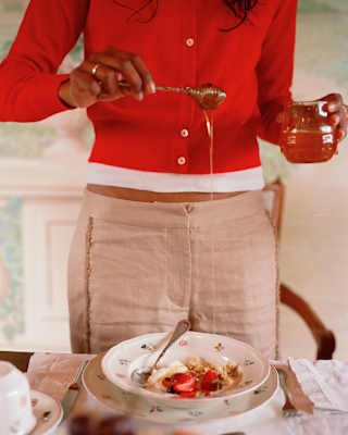 In close-up, a woman in a red cardigan and beige linen trousers holds a honey spool over a bowl of fruit, yoghurt and granola.