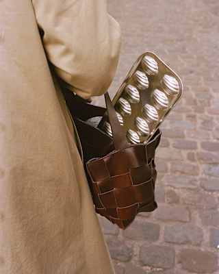 A silver-tone baking tray for making Madeleines juts from the leather bag of a woman in a beige coat, seen from behind.