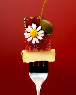 Artistic food image of an upright fork, prongs spearing a cube of cheese, topped with a fruit jelly, daisy flower, caper and gem.