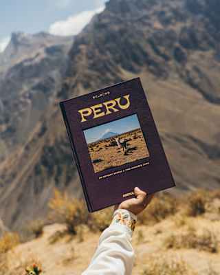 Against a mountain backdrop, a hand holds up a copy of Peru, with an alpaca cover-photo, created by Belmond with Assouline.