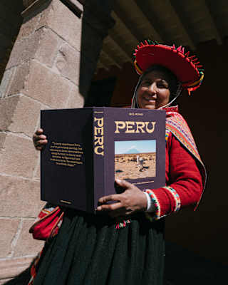 A Peruvian woman in brightly-woven traditional clothing holds open Belmond's travel book, Peru, with an alpaca cover-image.