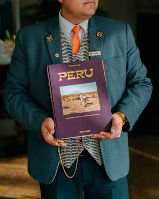 A man in a teal suit with gold hints and a patterned waistcoat holds a front-facing copy of Peru, by Belmond and Assouline.