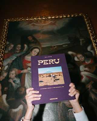 A woman holds up Peru, a book by Belmond and Assouline, obscuring her face, in front of an oil painting, seen at a low-angle.