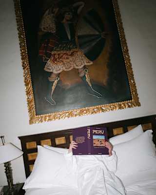 Tilted image of a woman in a white robe as she lies on a bed beneath an oil painting, reading up a copy of Belmond's book, Peru.
