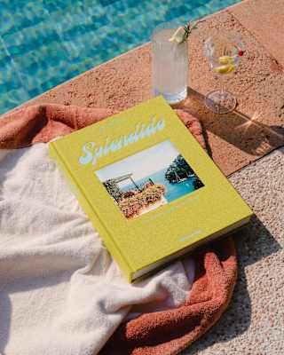 A copy of Splendido, with yellow binding and cover-photo of a bay view rests on a towel with two drinks by a glittering pool.