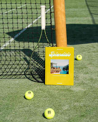 The Splendido book, bound yellow with a sea-view photo, leans against a tennis net post with a racquet and three loose balls.