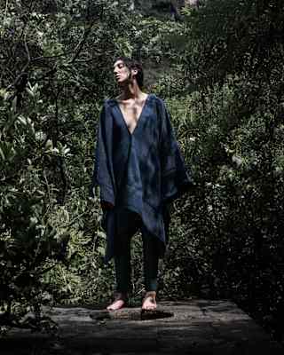 A dark-haired male model stands on a stone wall in dense gardens, in a black oversized V-neck top by Collectiva Concepción.