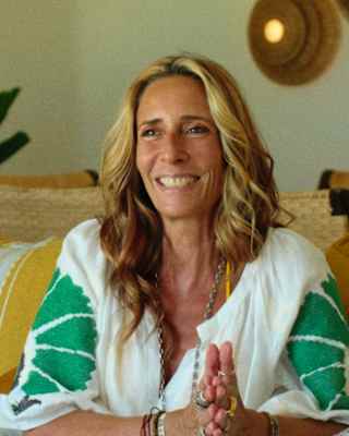 Tara Bernerd, with long blonde hair, smiles as she sits in a white and green top sitting on a sofa with earth-tone cushions.