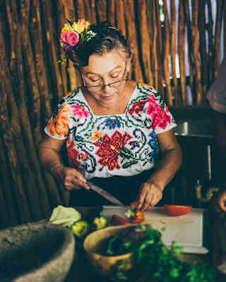 A woman wearing glasses and a colourful embroidered blouse slices vegetables in a kitchen. She has a flower in her hair, and there are various ingredients and a stone mortar on the table in front of her.