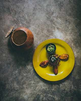 A yellow plate with three pieces of Indian-style fried food and a bowl of green chutney, placed on a grey surface next to a rustic brown clay pot with a dragonfly decoration.