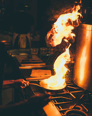 A person cooking at a hob creates a large burst of flame in a pan, illuminating the dim kitchen with orange and yellow light. Nearby, a large metal pot sits on the cooker.