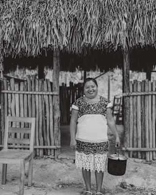 A woman wearing a traditional dress stands and smiles, holding a pot in front of a rustic wooden and thatched-roof building. A wooden chair is beside her. The photo is in black and white.