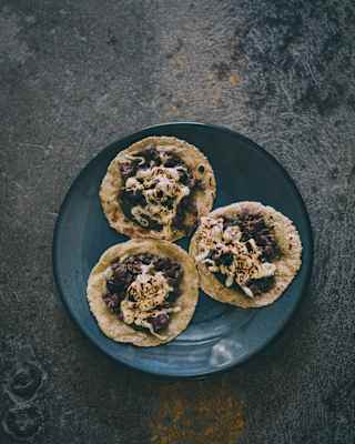 Three small tortillas topped with black beans and crumbled cheese are arranged on a dark plate, set against a textured, rustic surface.