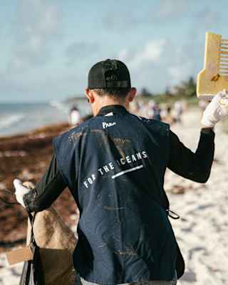 A person wearing gloves and a vest reading For the Oceans picks up rubbish on a beach, holding a large yellow plastic item and a sack, with other people cleaning in the background.