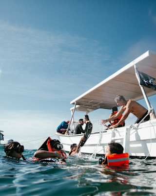 Four surfaced divers, two with buoyancy jackets, bob in the water between two boats of people who help their equipment on board.