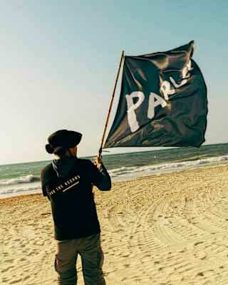 Person on a sandy beach holding a large black flag with the word PARLEY partially visible, facing the sea under a clear sky.