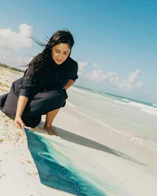 Artist Paola Dávila, with long dark hair and black clothing, creates a cyanotype turquoise and sand art piece on the beach.